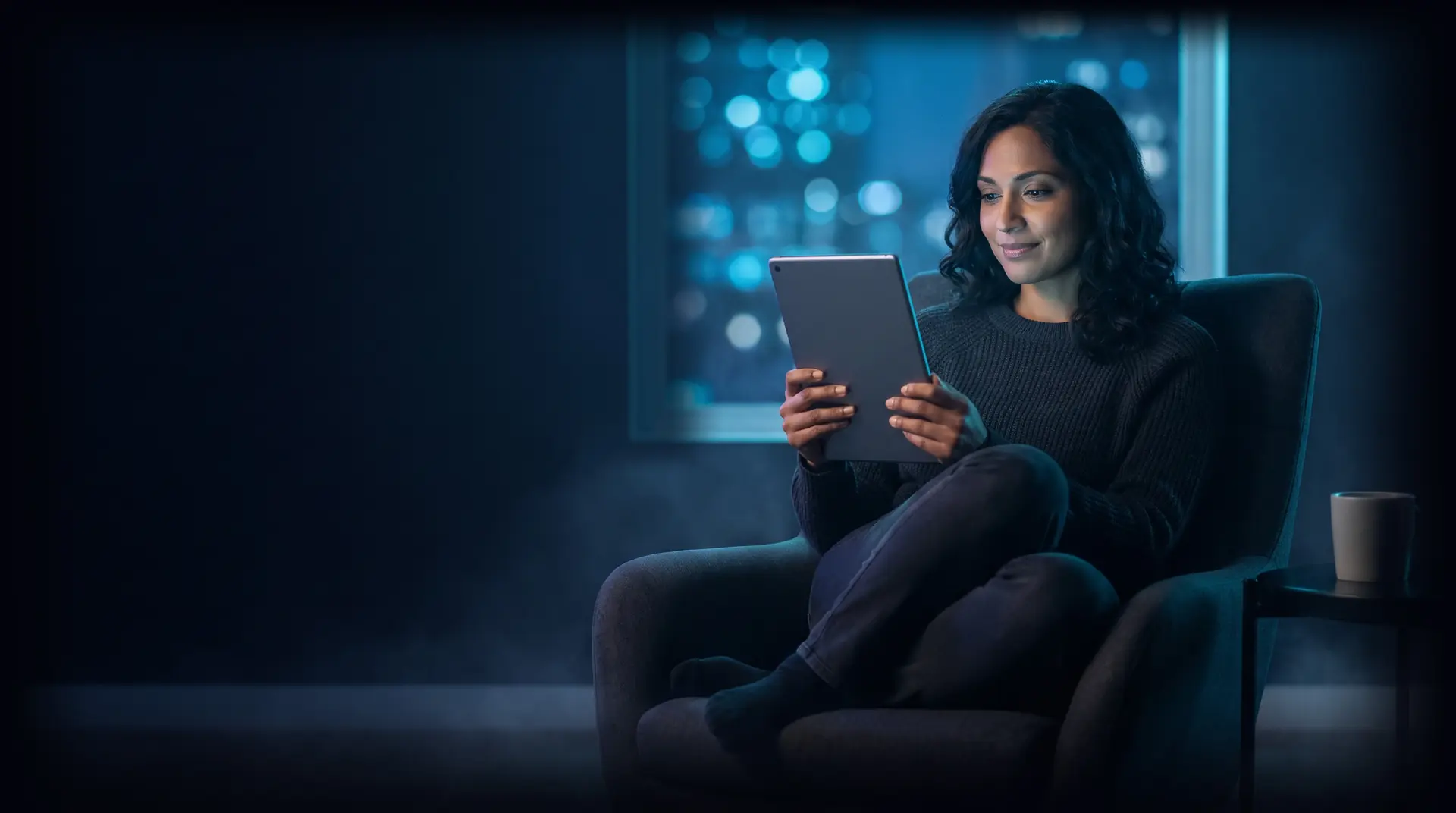 Mujer leyendo en una tableta de noche junto a un ventanal (imagen ilustrativa)